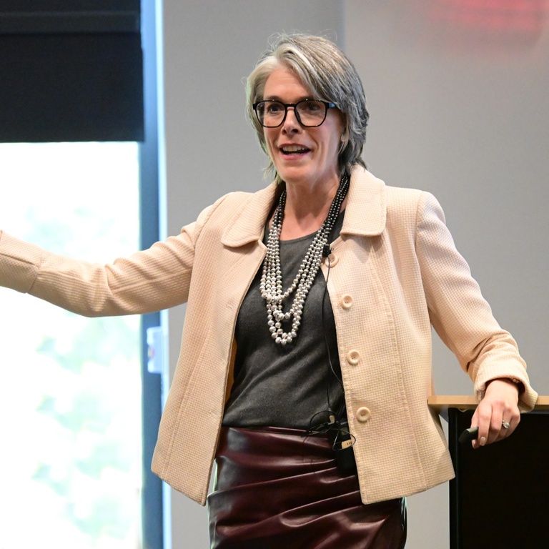 Susan Winckler speaking and gesturing with one hand. She is leaning against a podium.