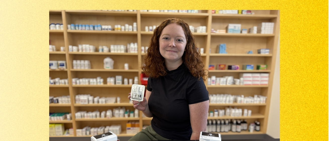 A University of Iowa College of Pharmacy PharmD student with curly red hair stands behind a pharmacy counter. She is holding a package of naloxone nasal spray. Naloxone boxes and informational sheets are displayed on the counter, with shelves of medications behind her. The photo is framed by a textured gold background.