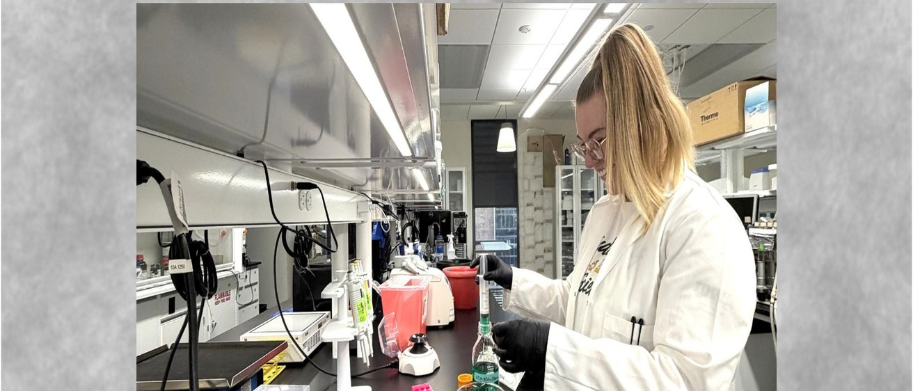 Researcher Sophie Granger, a member of University of Iowa College of Pharmacy Professor Michael Ashley Spies’ lab, is wearing a white lab coat and gloves. She is measuring a green liquid at a laboratory bench surrounded by beakers, pipettes, test tubes, and other research equipment.
