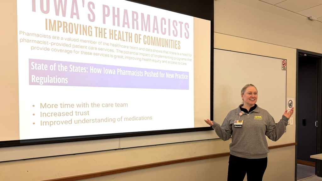 Presenter stands beside a projected slide reading “Iowa’s Pharmacists Improving the Health of Communities,” discussing new practice regulations. She gestures with both hands while speaking in a classroom setting. Slide highlights include increased trust, more time with the care team, and improved understanding of medications.