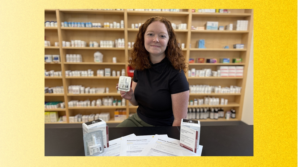 A University of Iowa College of Pharmacy PharmD student with curly red hair stands behind a pharmacy counter. She is holding a package of naloxone nasal spray. Naloxone boxes and informational sheets are displayed on the counter, with shelves of medications behind her. The photo is framed by a textured gold background.